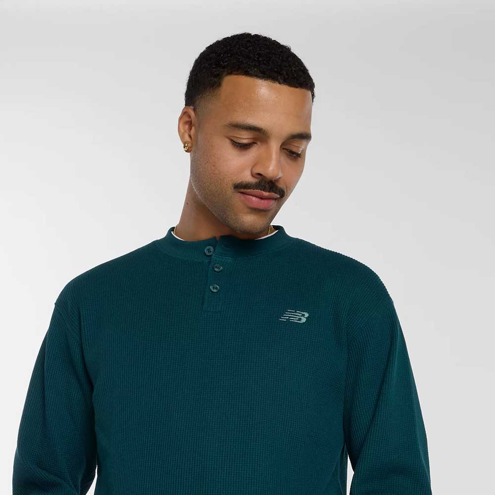 A man with short curly hair and a mustache, smiling softly, wears a teal New Balance Numeric Waffle Knit Tee MT51958AAV. The cotton-blend shirt features the logo on the left chest. The background is plain and light-colored.