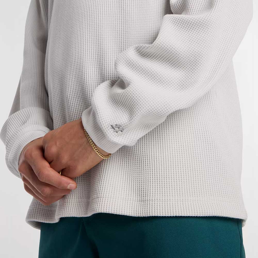 Wearing the New Balance Numeric Waffle Knit Tee MT51958GYM and teal pants, a person stands with their hands gently clasped, accessorized with a gold chain bracelet.