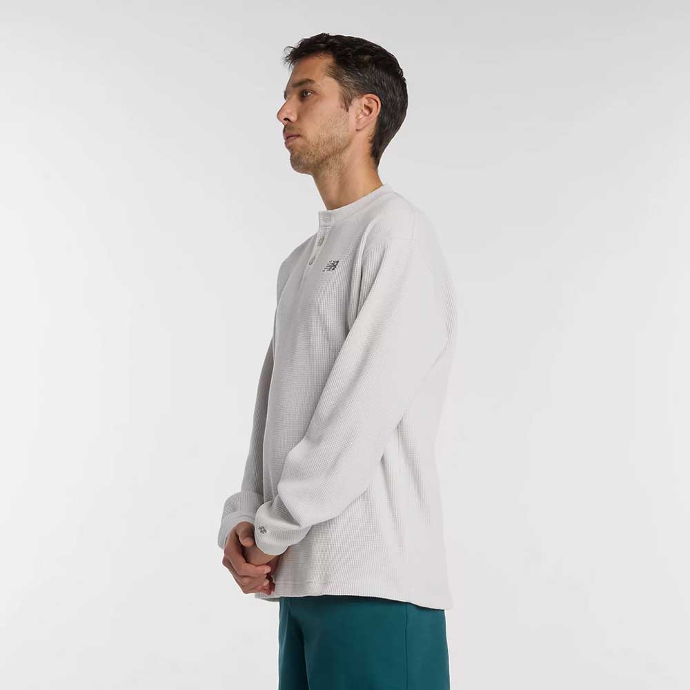A man stands in profile against a white background, wearing the New Balance Numeric Waffle Knit Tee MT51958GYM in light gray and teal pants. His hands are clasped in front of him as he looks straight ahead.