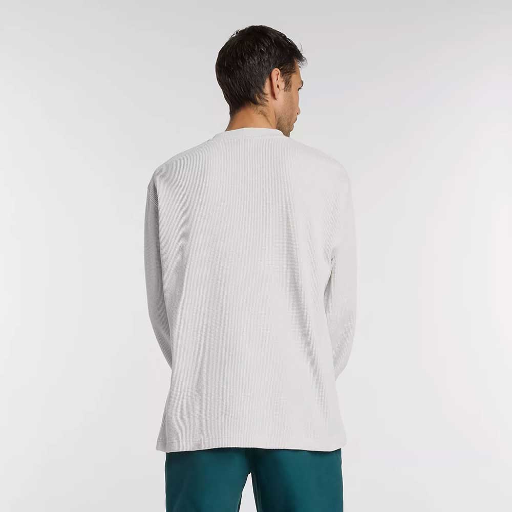 A man with short dark hair faces away from the camera, wearing a New Balance Numeric Waffle Knit Tee MT51958GYM in light gray and teal pants, standing against a plain white background.