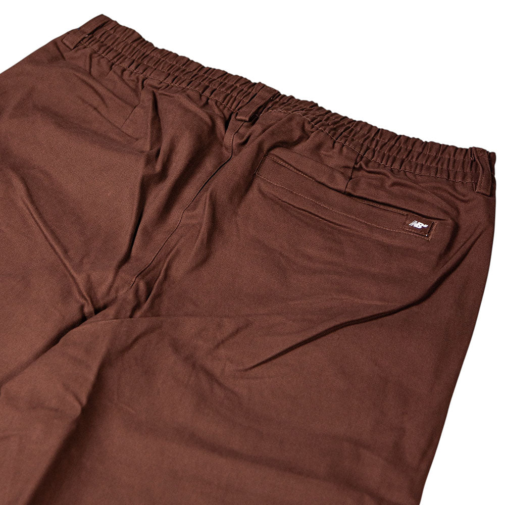 The New Balance Numeric Pant Pumperknickel MP51953ABJ features brown cotton canvas, an elastic waistband, a rear pocket with a white tag, and a relaxed straight-leg fit—ideal as everyday skateboarding pants. Laid flat on a white background.