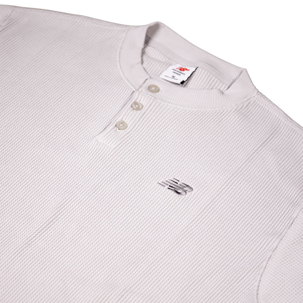 A close-up of the New Balance Numeric Waffle Knit Tee MT51958GYM in light gray, featuring long sleeves, waffle-knit texture, two buttons, an embroidered New Balance logo on the chest, ribbed collar, and a branded neckline tag.