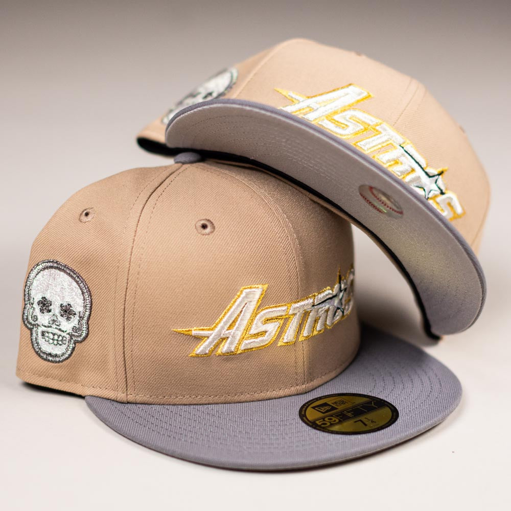 Two beige-and-gray Houston Astros New Era hats: one features a Chalino Skull patch and the other has an Astros logo under the brim. Both Camel Storm 5950 fitted caps boast striking gold and white embroidery.