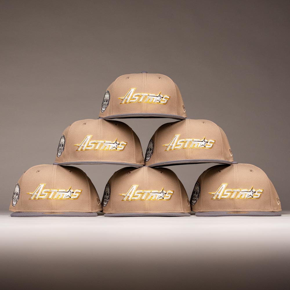 Six Chalino Skull Camel Storm Houston Astros New Era Hat 5950 Fitted caps are stacked in a pyramid on a light surface, each showing “Astros” in gold. The neutral gray gradient background highlights their sleek design.