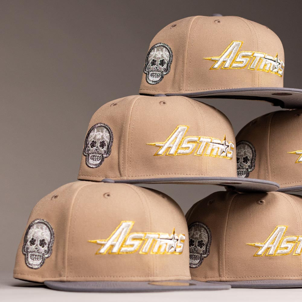 A stack of Chalino Skull Camel Storm Houston Astros New Era Hat 5950 Fitted, featuring embroidered skull designs and "Astros" in gold and white on the front, arranged in a pyramid against a neutral background.