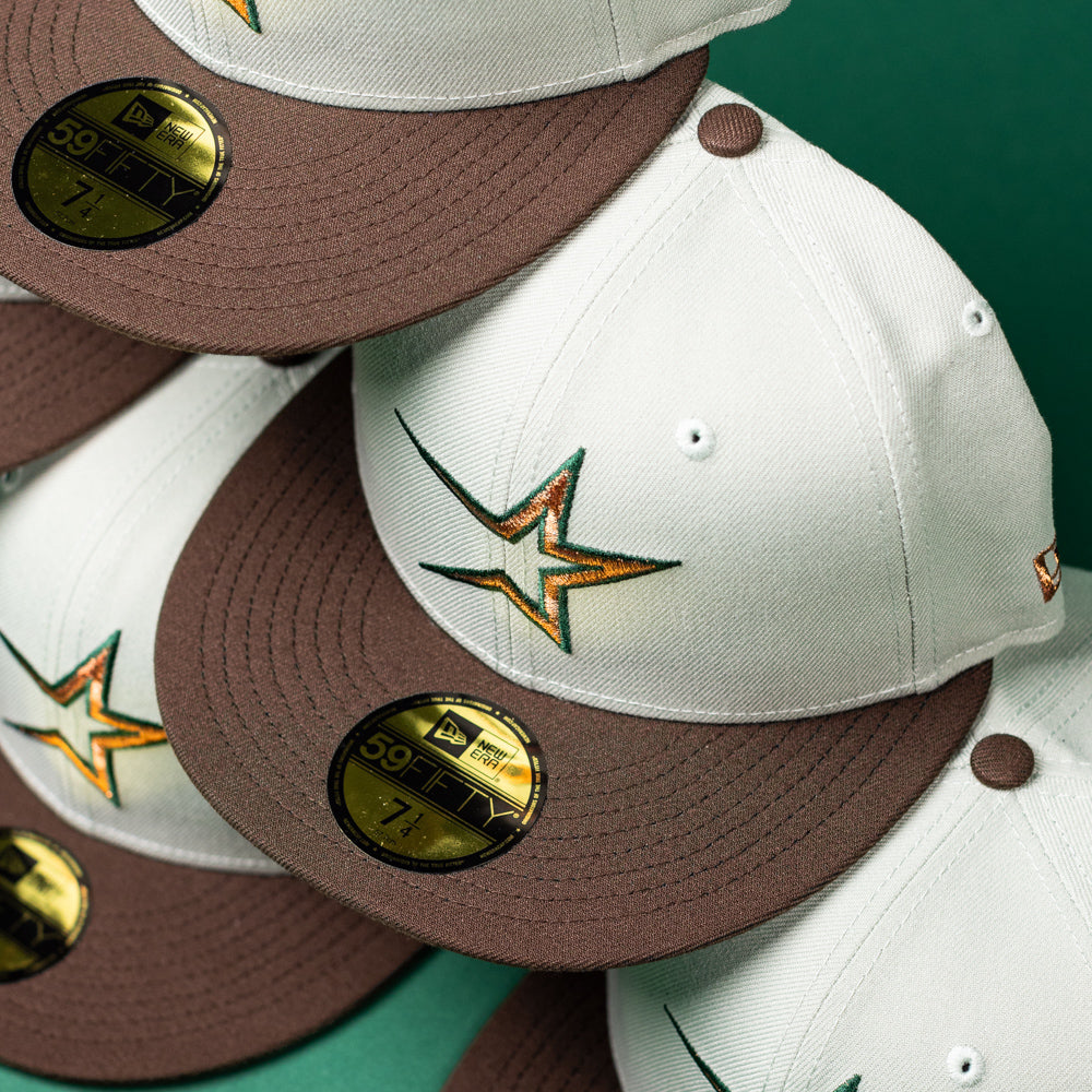 A stack of Copper Trails OG Walnut Egreen Houston Astros New Era Hat 5950 Fitted caps, featuring the team’s embroidered star logo and gold 59FIFTY New Era stickers, are arranged diagonally on a green background.