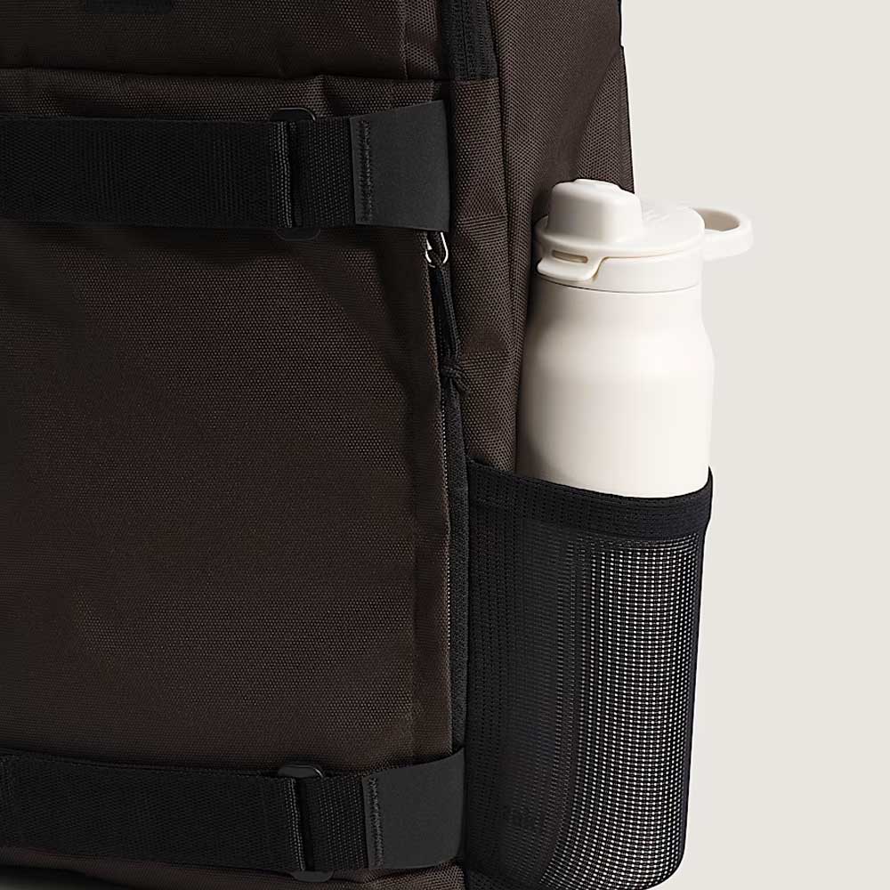 A white water bottle rests in the black mesh side pocket of a dark brown Obstacle Skatepack Backpack VN0A3I69OVB1 Brown Ripstop from Vans Skateboarding, set against a plain light background.