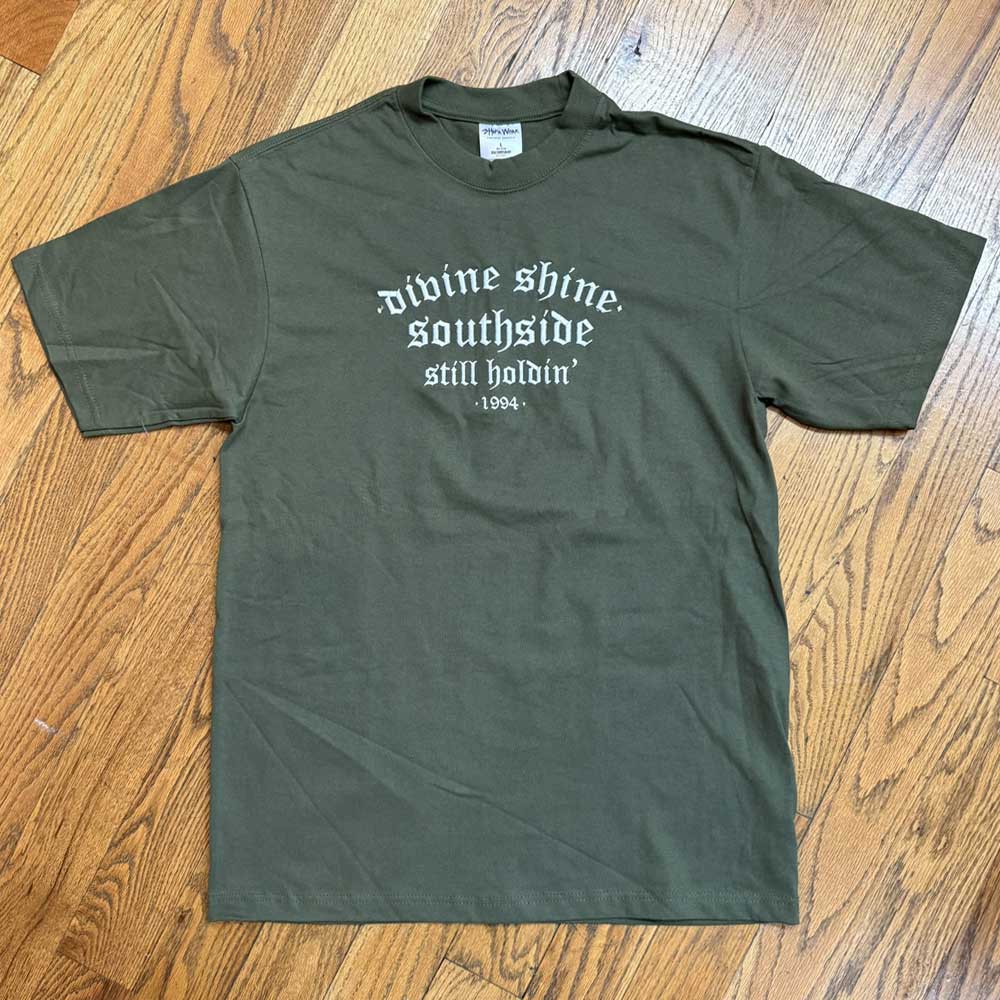 The Olive Divine Shine x Southside Still Holdin Max Heavyweight Tee, inspired by Southside Skatepark, lies flat on a wooden floor and features "divine shine. southside still holdin 1904." embroidered in white on the front.