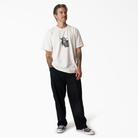 A tattooed man in a white graphic tee, black sneakers, and P415314 Mike Anderson Black Twill Pant Dickies Skateboarding stands against a plain white background, looking slightly to the side with one hand in his pocket.