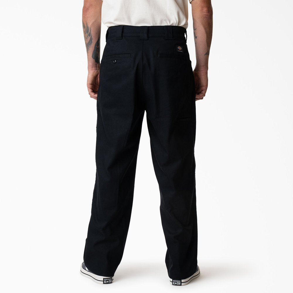 A person stands facing away, wearing black-and-white sneakers and P415314 Mike Anderson Black Twill Pant Dickies Skateboarding. The durable pants have flex fabric, belt loops, pockets, and a small circular logo above the back right pocket. White background.