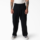 A person stands facing forward against a plain white background, wearing loose-fitting P415314 Mike Anderson Black Twill Pant Dickies Skateboarding, a white shirt, and black-and-white sneakers. Their tattooed arms hang by their sides.