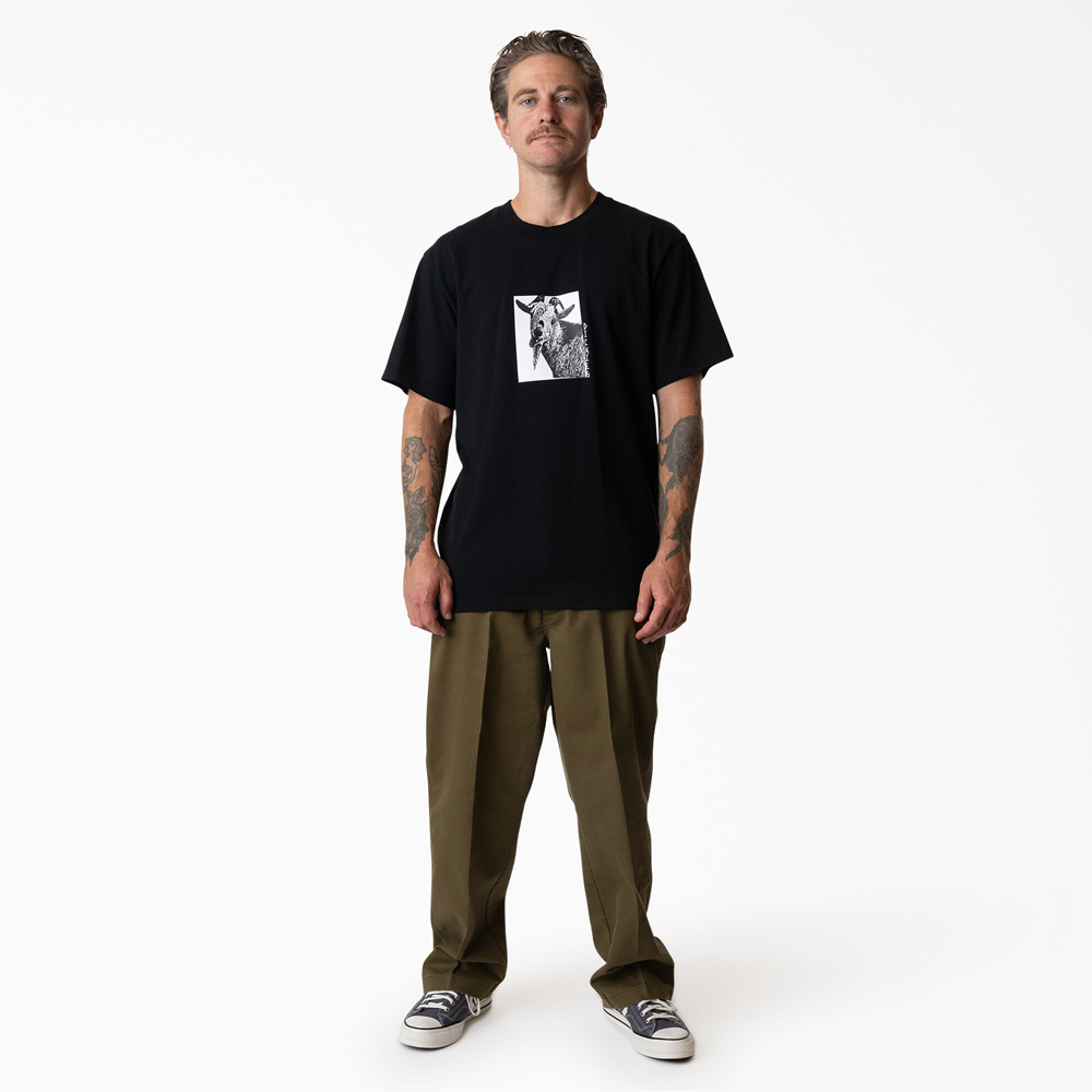 A man with tattooed forearms stands facing forward, wearing the P415867 Mike Anderson Goat Black Tee from Dickies Skateboarding, paired with olive green pants and black-and-white sneakers. The background is plain white.