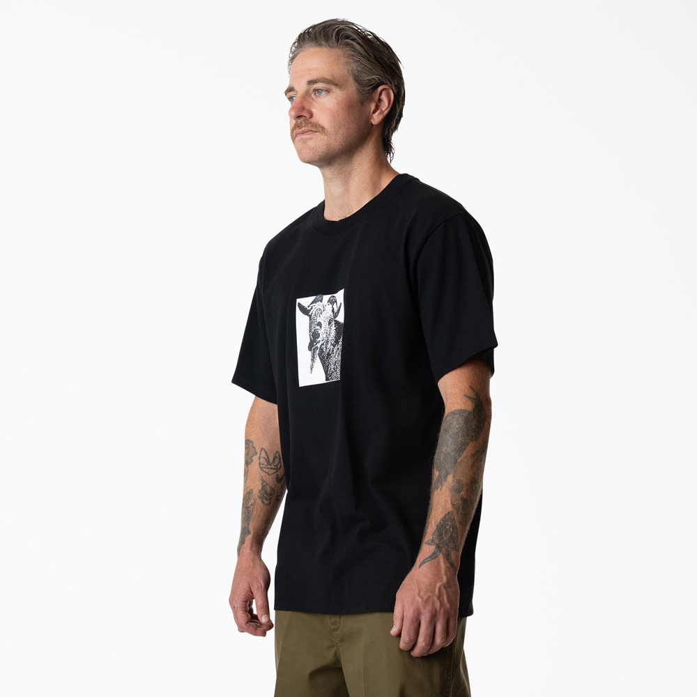 A man with tattooed arms wears the Dickies Skateboarding Mike Anderson Goat Black Tee (P415867) with a graphic design and cooling finish, paired with khaki pants, standing against a plain white background.