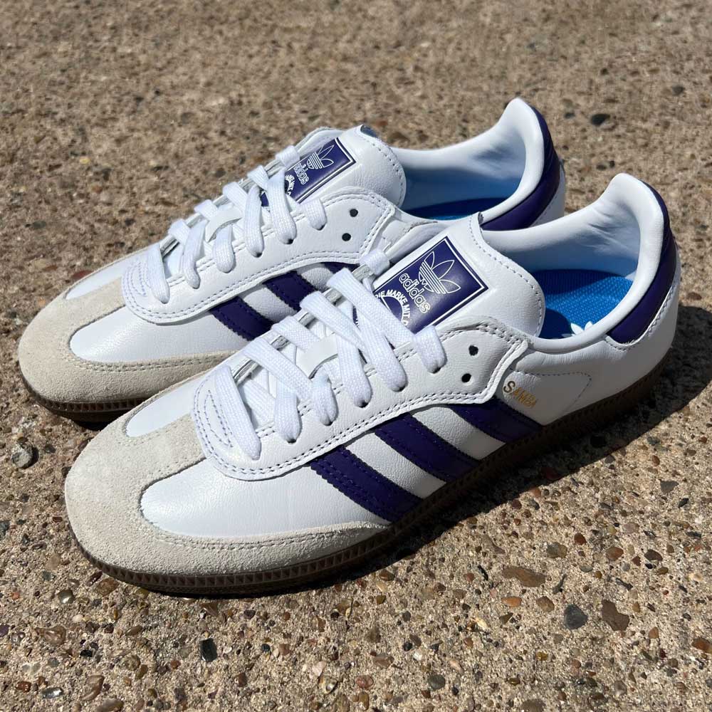 A pair of purple and white Samba ADV Adidas Skateboarding leather suede shoes rest on a sunlit textured concrete surface, featuring gold Samba ADV text on the side.
