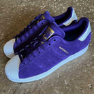 The Purple Zero Gold Superstar ADV Adidas Skateboarding Suede Shoes feature white shell toes and heels, reflective badging, and "SUPERSTAR" on the side—ideal for Adidas Skateboarding fans. Placed on textured concrete.