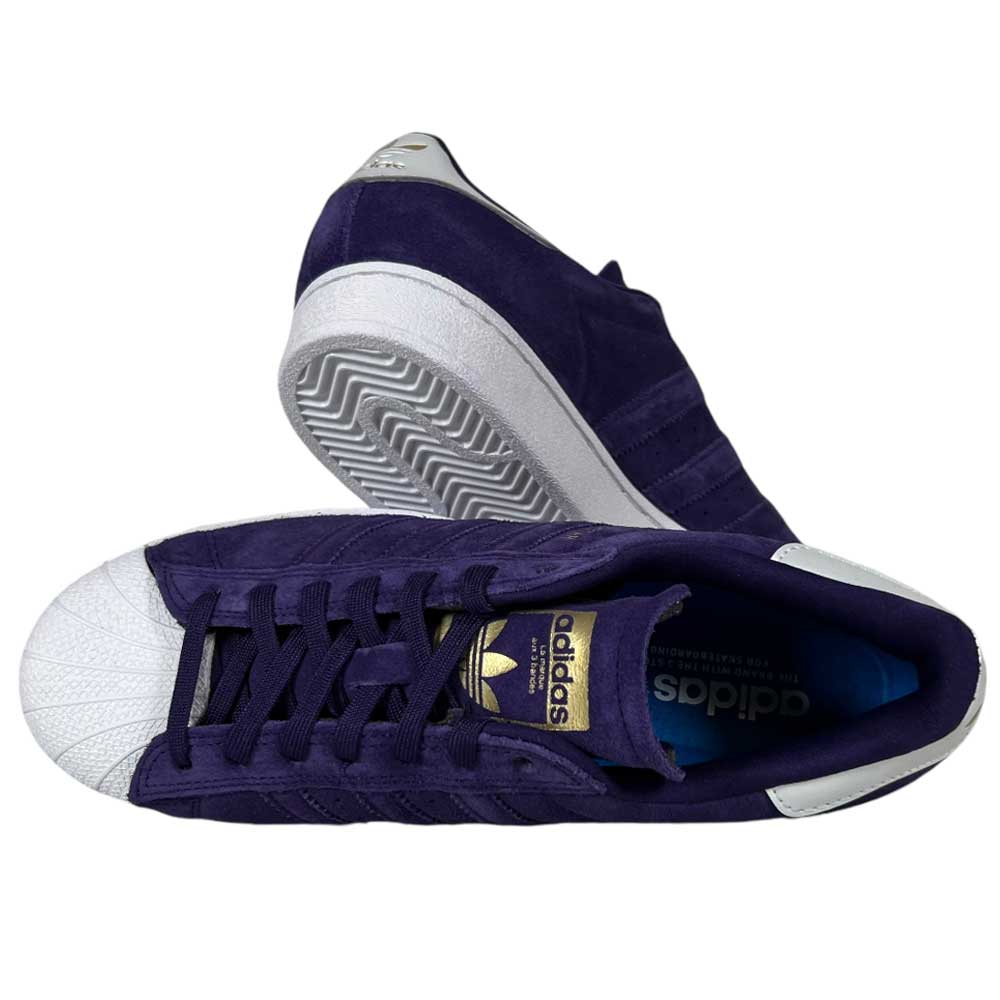 A pair of Purple Zero Gold Superstar ADV Adidas Skateboarding suede shoes with white shell toes, reflective badging, and logo. One shoe is upright showing laces; the other lies sideways, revealing the sole and design features.
