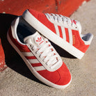 A pair of JP5861 Red White Gold Gazelle ADV Adidas Skateboarding suede shoes is placed on a concrete surface near a red-painted curb.
