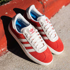 A pair of JP5861 Red White Gold Gazelle ADV Adidas Skateboarding Suede Shoes with white laces and blue insoles rests on concrete beside a red curb in bright sunlight.