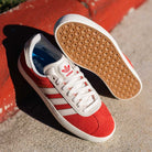 A pair of JP5861 Red White Gold Gazelle ADV Adidas Skateboarding suede shoes with gum soles and three white stripes sits on concrete beside a red curb.