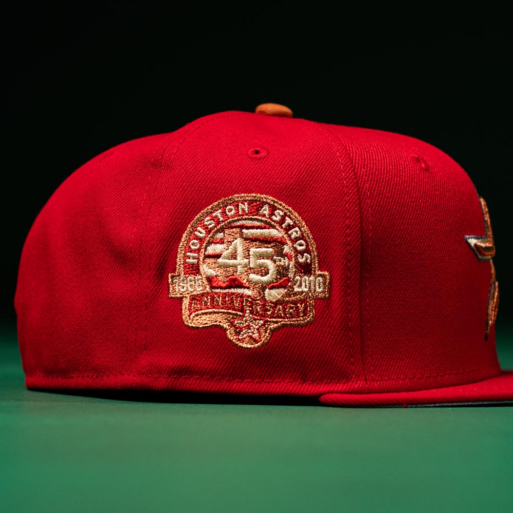 A Rusty Screw OG Scarlet Copper Houston Astros New Era 5950 Fitted hat rests on a green surface against a dark background, featuring scarlet and copper tones with a special embroidered patch celebrating the team’s 45th anniversary (1965-2010) on the side.