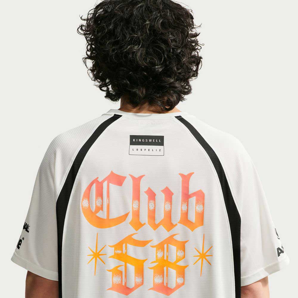 A person with curly hair is seen from behind, wearing the Nike SB Skateboarding Sail Black IR6760133 Skate Jersey—a white polyester shirt with black details, large orange "Club 58" text, and small Kingswell and Los Feliz labels near the neck.