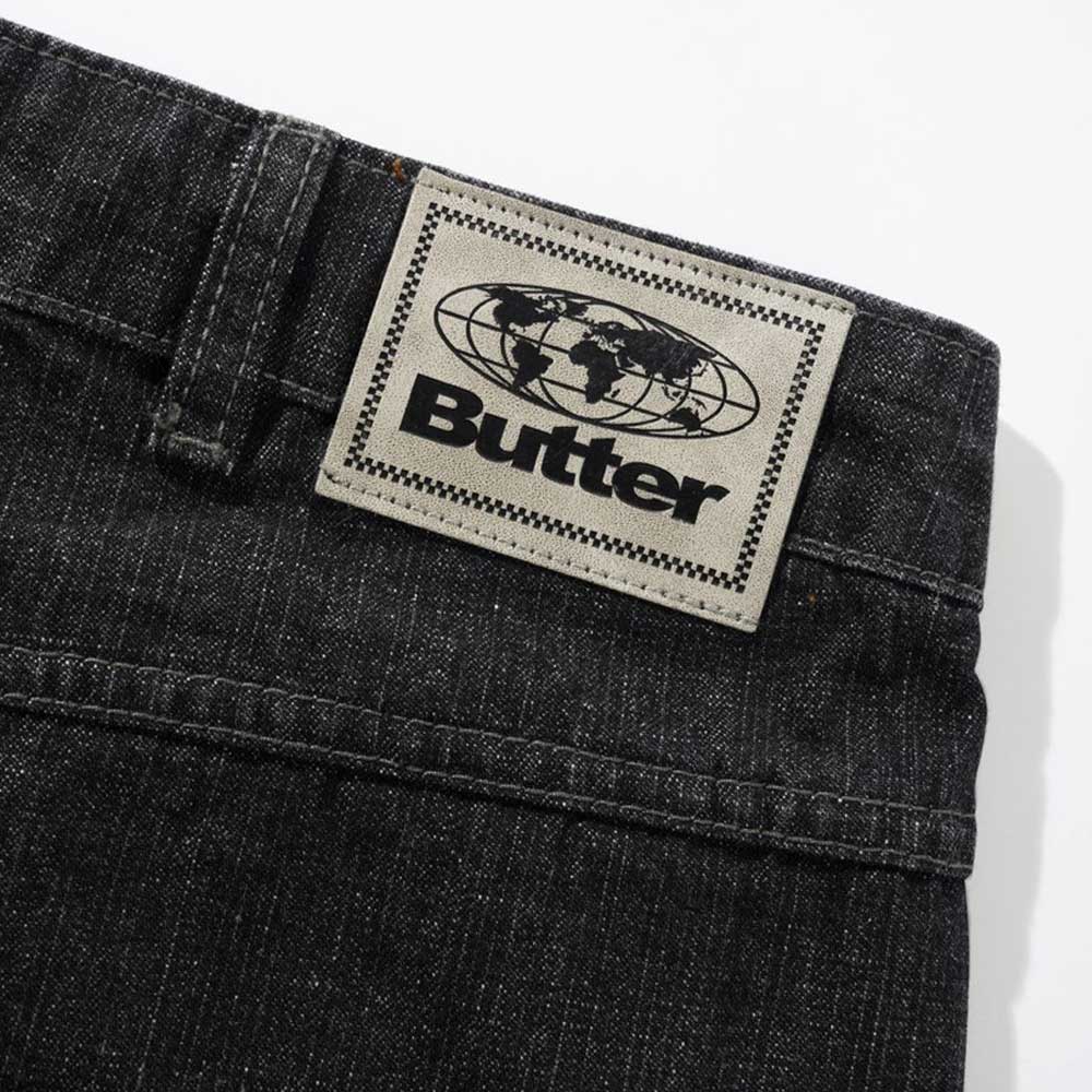 Close-up of Distressed Flat Black Relaxed Denim Jeans by Butter Goods, made from 100% cotton, featuring a beige “Butter” patch with globe graphic on the waistband.