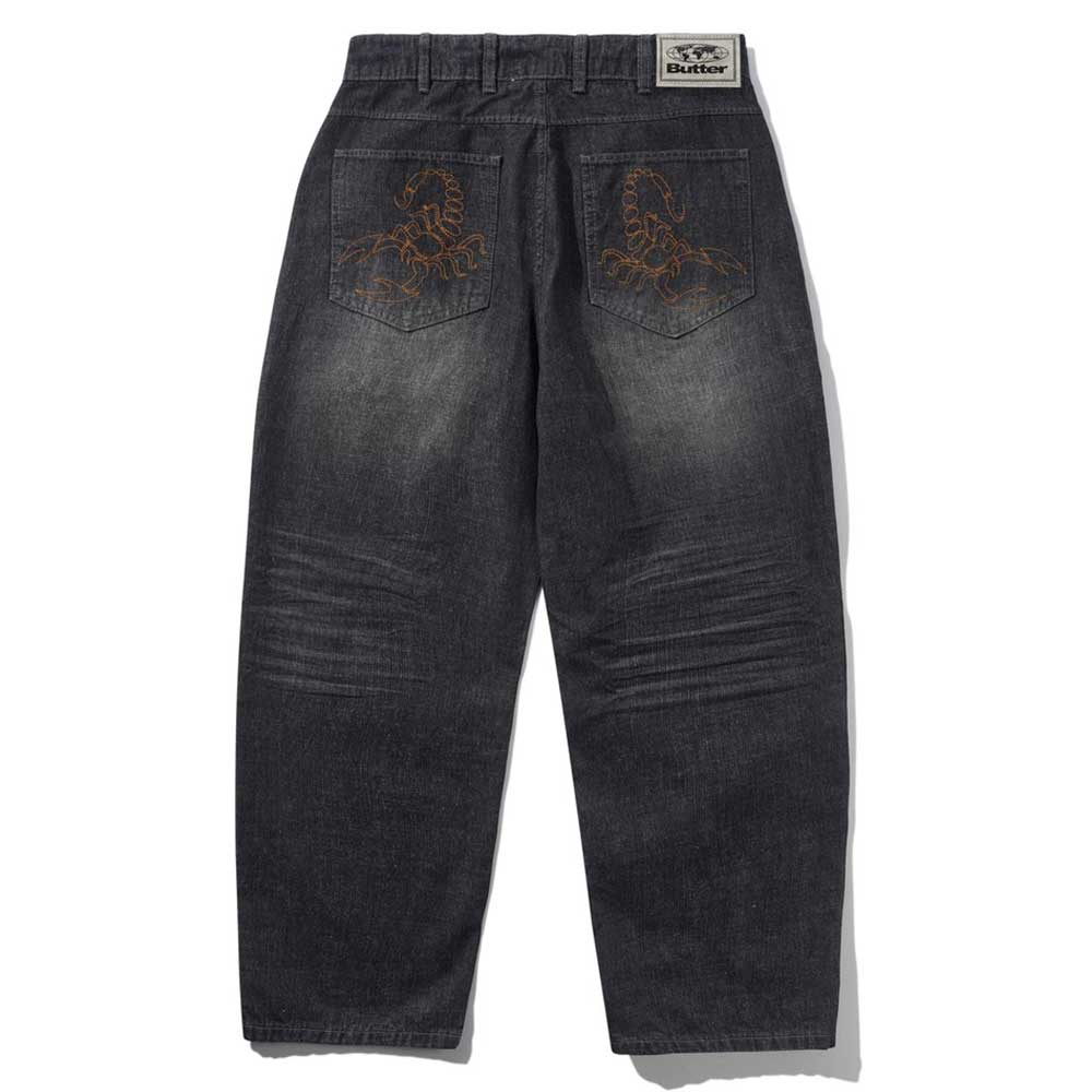 The Distressed Flat Black Relaxed Denim Jeans by Butter Goods feature a faded black finish, back embroidered orange scorpions, a white "Butter" label on the waistband, and are crafted from 100% cotton for comfort.