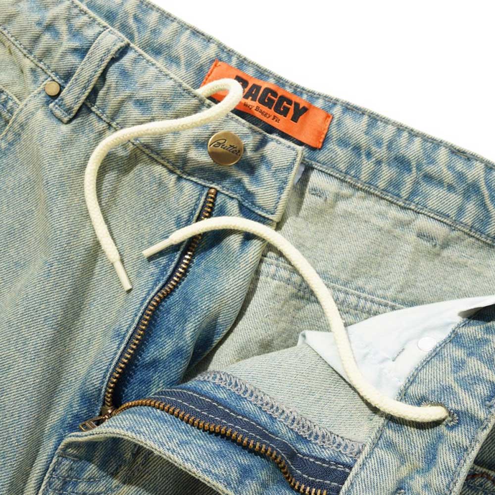 Close-up of Scorpion Stitch Tinted Light Blue Denim Jeans by Butter Goods, featuring a baggy fit, open zipper, white drawstring, orange BAGGY label inside the waistband, and embroidered patch pockets.