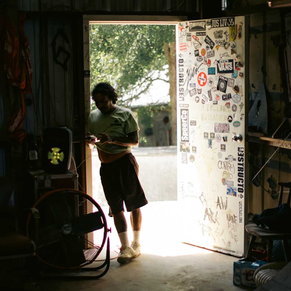 Wearing the Sebo Walker x Southside Skatepark Avocado Tee, a person stands in a sticker-covered, dim room, sunlight illuminating the entrance as they examine something in their hands—reminiscent of a Southside Skatepark scene.