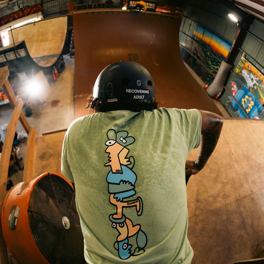 Wearing the Sebo Walker x Southside Skatepark Avacado Tee, a skateboarder stands helmeted at the edge of Southside Skatepark’s indoor ramp, ready to drop in, with bold graffiti and energy filling the scene.
