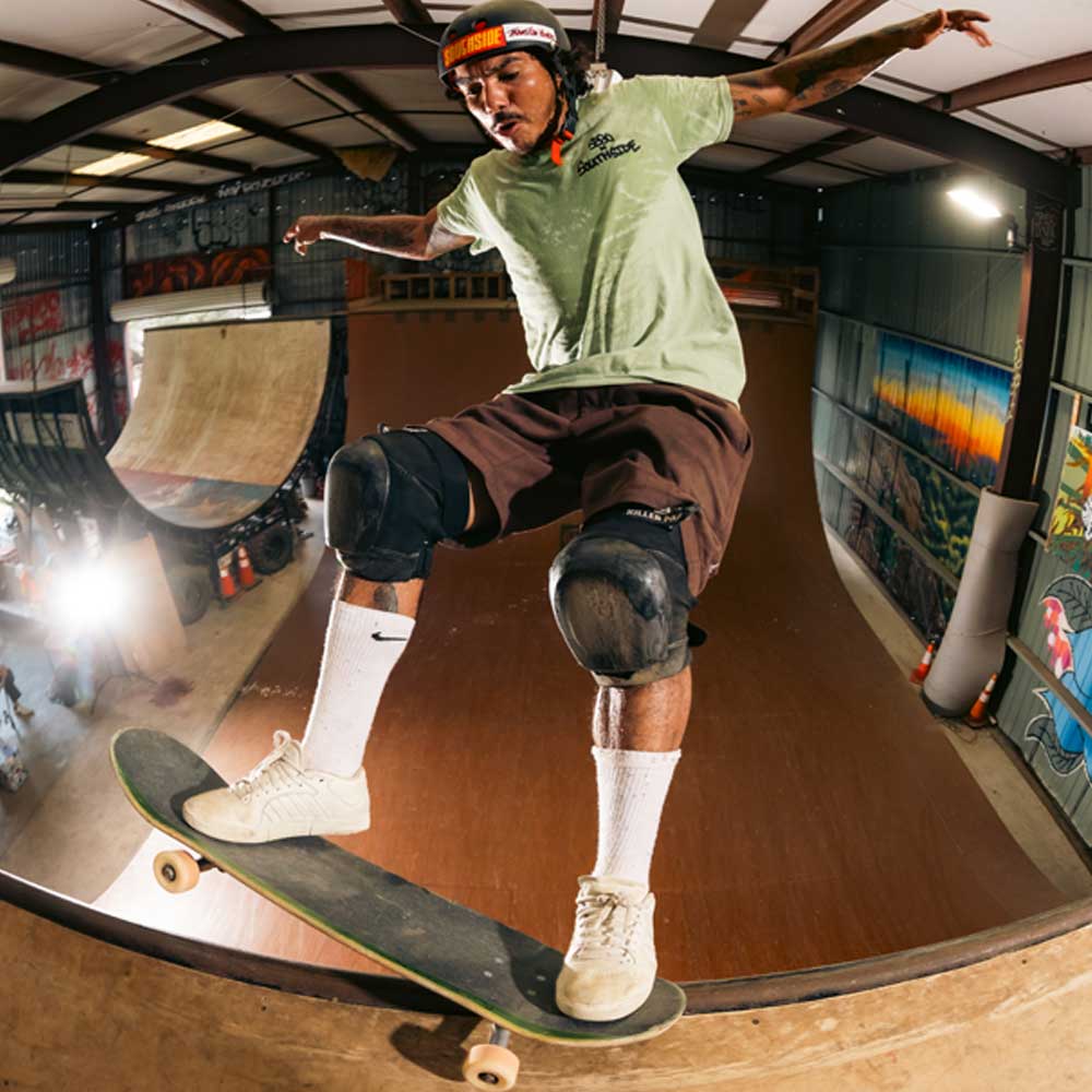 Wearing the Sebo Walker x Southside Skatepark Avocado Tee, a skateboarder in a helmet and knee pads performs a trick on the edge of an indoor ramp at Southside Skatepark, with graffiti art lighting up the walls.