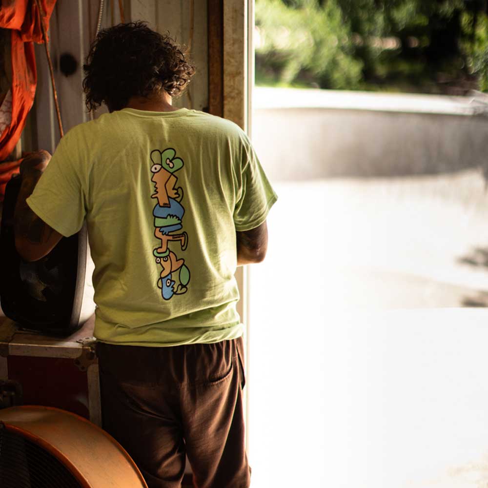 A person with curly hair stands indoors near an open door at Southside Skatepark, wearing the Sebo Walker x Southside Skatepark Avocado Tee featuring colorful abstract art on the back as sunlight streams in, brightening the scene.