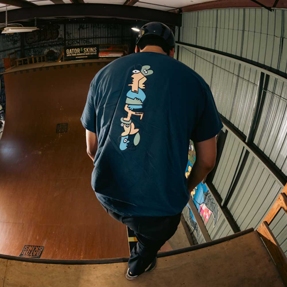 Wearing the Sebo Walker x Southside Skatepark Blueberry Tee, a person stands at the edge of Southside Skatepark’s indoor ramp, facing away; the blue tee displays a vertical design of colorful, abstract cartoon faces and a matching cap.