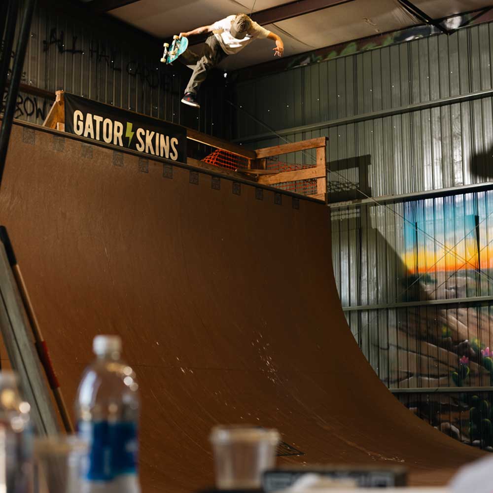 A skateboarder, likely Sebo Walker, soars above a half-pipe at Southside Skatepark wearing the Sebo Walker x Southside Skatepark Oat Tee, with Gator Skins signage and blurred bottles visible in the warehouse backdrop.
