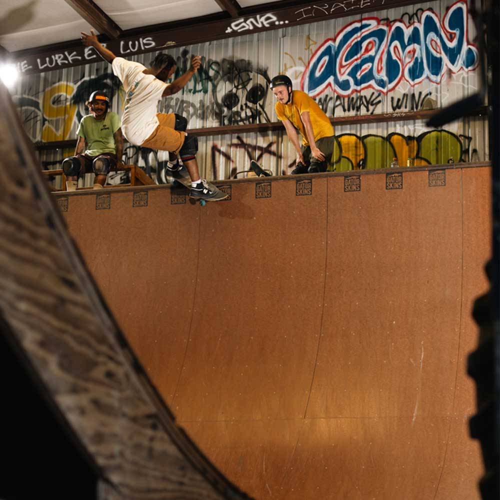 A person skateboards on an indoor half-pipe at Southside Skatepark, wearing the Sebo Walker x Southside Skatepark Oat Tee and protective gear, while two others watch from the top against a backdrop of colorful graffiti.