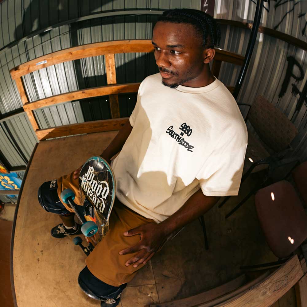 A man sits on a bench at Southside Skatepark, indoors near corrugated metal walls and wooden railings, holding a skateboard and wearing the Sebo Walker x Southside Skatepark Oat Tee with brown shorts.