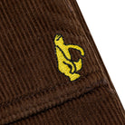 Close-up of Shmoo Carpenter Brown Corduroy Krooked Pant fabric featuring a small, embroidered yellow Shmoo logo stitched on the ribbed, textured material, highlighting the unique Krooked Pant style.