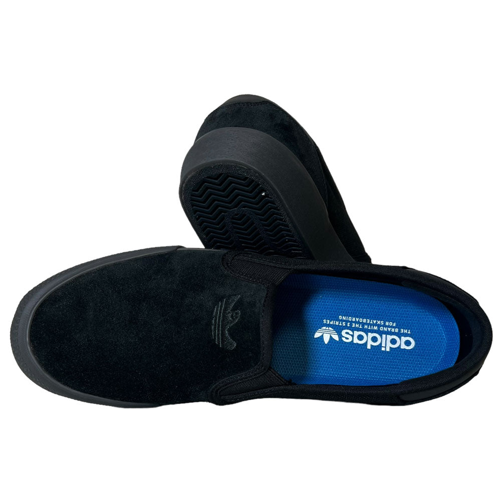 A pair of Shmoofoil Slip Adidas Skateboarding Core Black Carbon Suede Shoes with blue insoles, shown from a top and side angle. The Adidas logo appears on the upper and insole; patterned grip soles are visible.