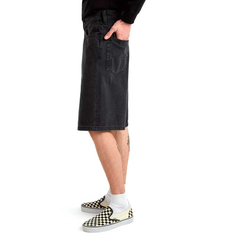 Wearing Skate Check 5 Loose Shorts Black Denim Vans Skateboarding, white socks, and black-and-white checkered slip-ons, a person stands waist-down side view, one hand in pocket.