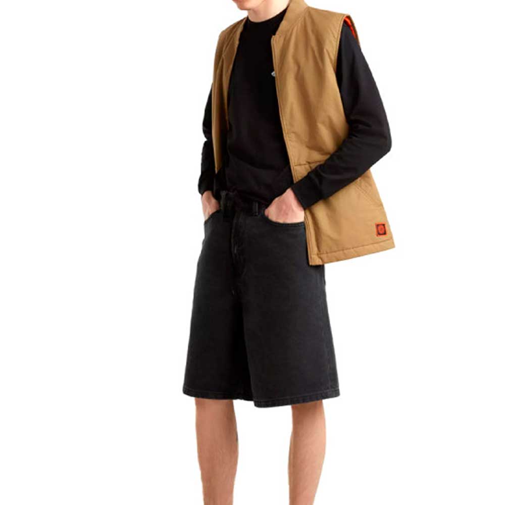 A person stands with hands in pockets, wearing a tan vest over a black long-sleeve shirt and Vans Skateboarding Skate Check 5 Loose Shorts in black denim. The individual faces slightly to the side, and their head is not visible in the image.