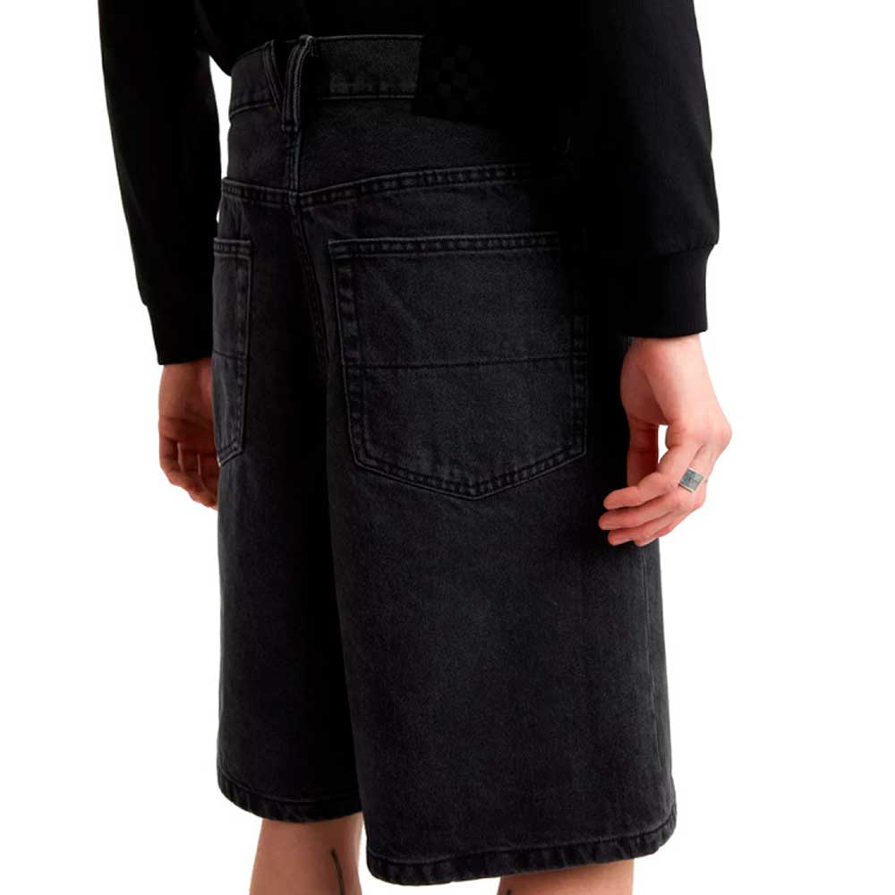 A person wearing a black long-sleeve shirt and Vans Skateboarding Skate Check 5 Loose Shorts in black denim stands facing away, highlighting the loose fit, large back pockets, and a ring on their right hand.