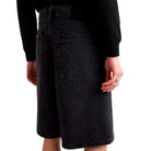A person wearing a black long-sleeve shirt and Vans Skateboarding Skate Check 5 Loose Shorts in black denim stands facing away, highlighting the loose fit, large back pockets, and a ring on their right hand.