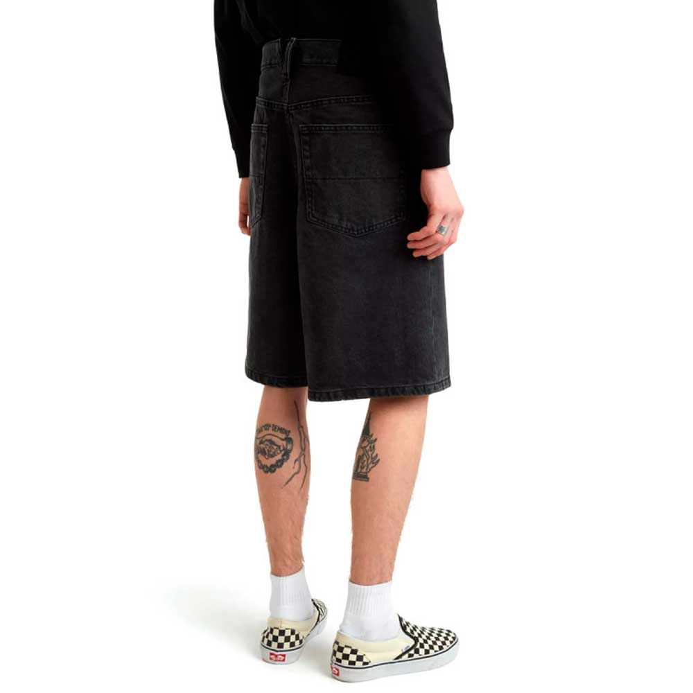 A person stands with their back to the camera, wearing a black sweatshirt, Vans Skateboarding Skate Check 5 Loose Shorts in black denim, white socks, checkered slip-on shoes, and displaying tattoos on the backs of both legs.