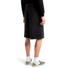 A person stands with their back to the camera, wearing a black sweatshirt, Vans Skateboarding Skate Check 5 Loose Shorts in black denim, white socks, checkered slip-on shoes, and displaying tattoos on the backs of both legs.