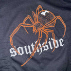 The Southside Tee Spider Black Orange Reflective features an orange spider with a white mark above bold, gothic-style “southside” lettering on dark 100% USA cotton—streetwear style.