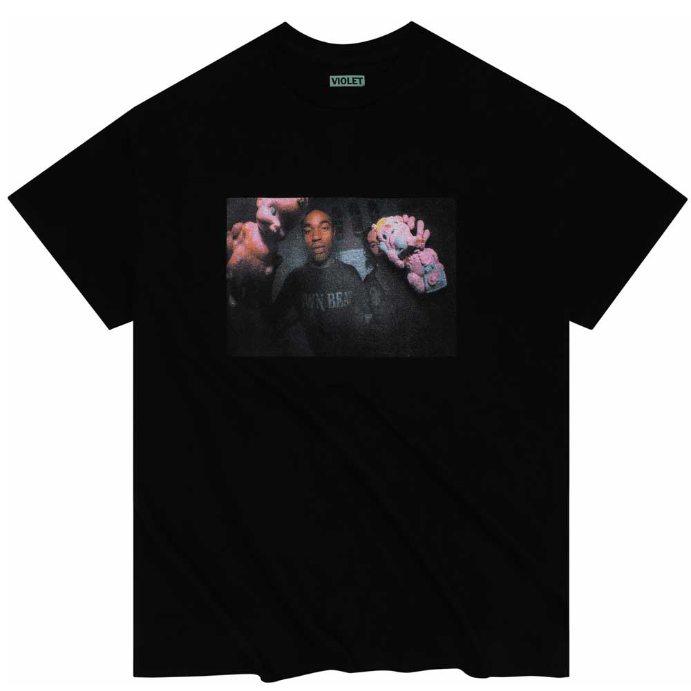 The Squeaky Violet Skateboards Black Tee is a black T-shirt featuring a slightly grainy, rectangular graphic of a person holding two plush toys—one pink and one purple—centered on the chest. The shirt includes VIOLET branding on the neck tag and is part of the Violet Skateboards collection, reflecting William Strobeck’s signature visual style.