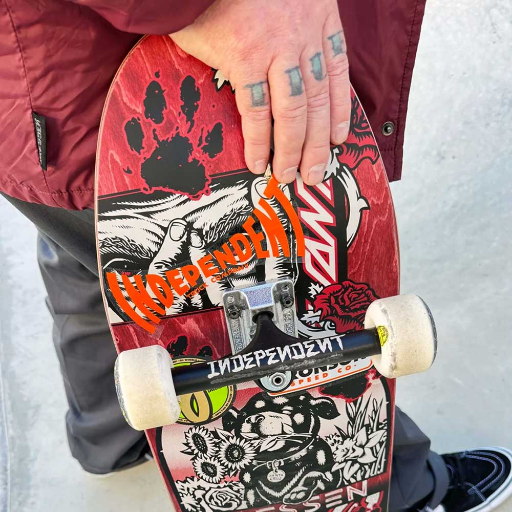 A person with tattoos on their fingers holds a skateboard covered in colorful stickers, including Independent logos, while wearing a maroon jacket, dark pants, and Stage 4 Eric Dressen Pro 151 Black Silver Standard INDY Trucks.