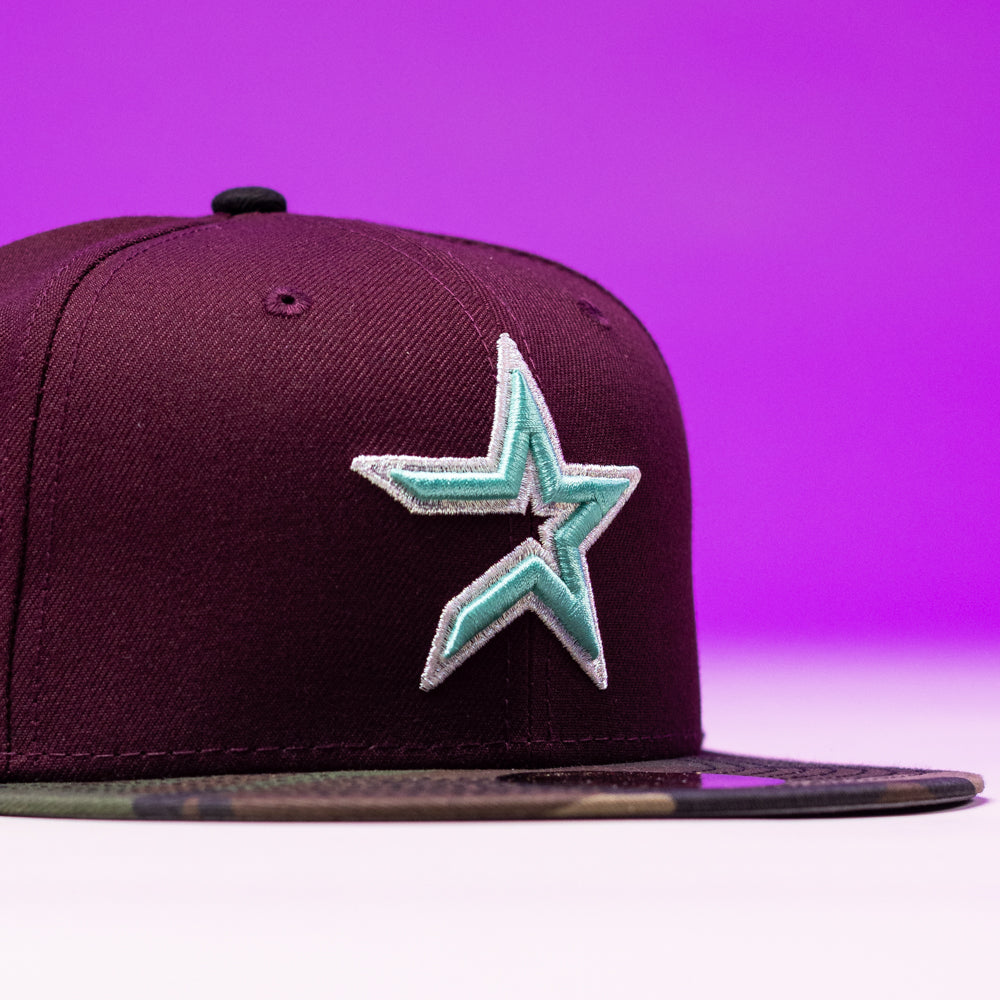 The Swag Era 50th Camo Maroon Houston Astros New Era Hat 5950 Fitted features a teal and white embroidered star logo on the front and a camouflage-patterned flat brim, presented against a purple background.