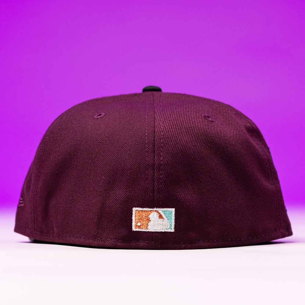 A maroon Swag Era 50th Camo Houston Astros New Era 5950 Fitted Hat is shown from the back, with a small rectangular MLB logo patch near the bottom, set against a vibrant purple background.