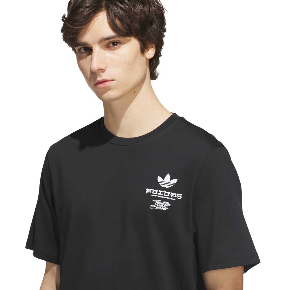 A young person with short brown hair wears the TOS Crane Tee Black White KS4336 from Adidas Skateboarding, a black t-shirt featuring bold white graphics and text—including the Adidas logo—suggesting a collaboration with Temple of Skate. They stand against a plain background, gazing slightly to the side.